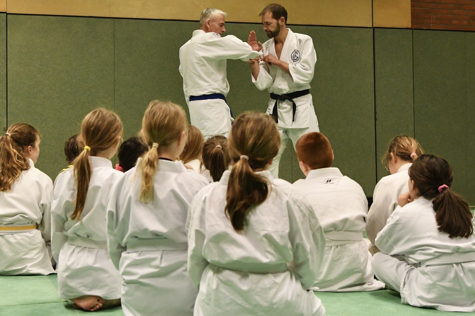 Zwei Judoka demonstrieren eine Technik, w&auml;hrend eine Gruppe von Kindern im Hintergrund zuschaut.