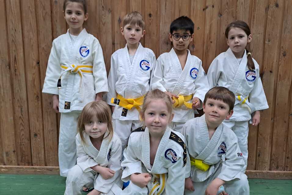 Gruppe von sieben Kindern in wei&szlig;en Karateanz&uuml;gen mit gelben G&uuml;rteln, l&auml;chelnd vor einer Holzwand.