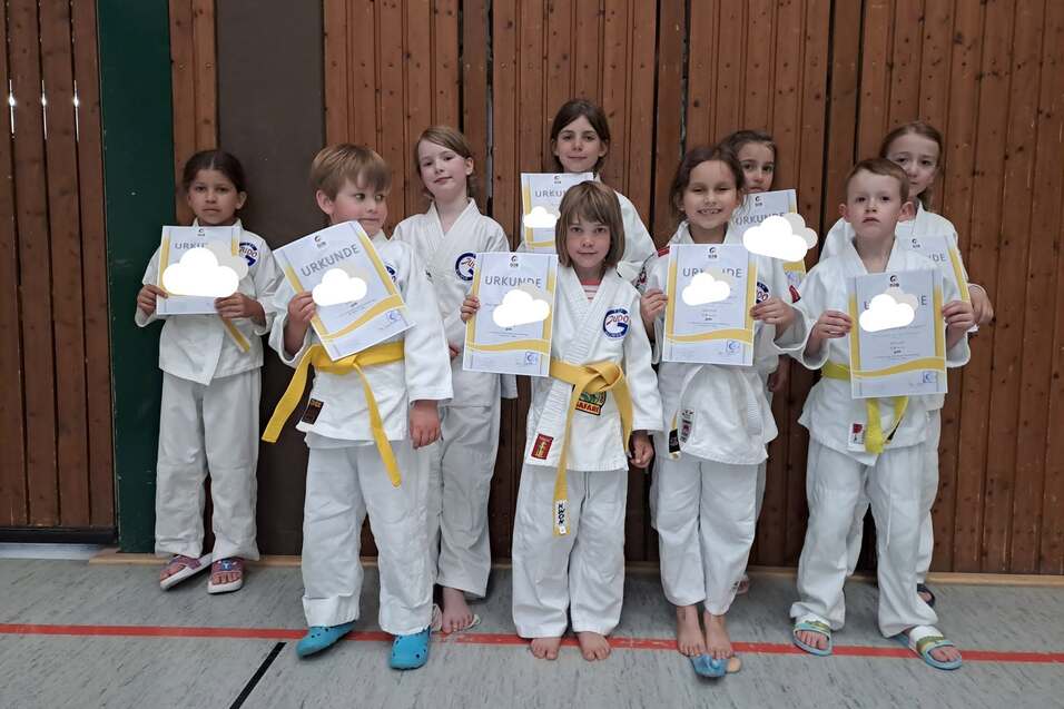 Gruppe von Kindern in wei&szlig;en Judo-Anz&uuml;gen mit gelben G&uuml;rtel, alle halten Urkunden in einer Sporthalle.