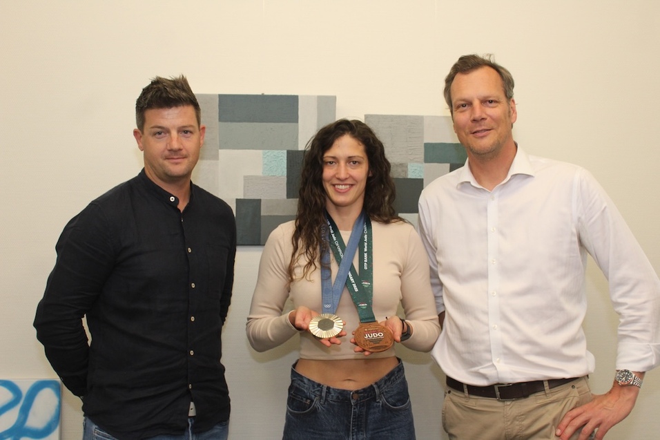 Drei Personen posieren vor einer Kunstwand, wobei eine Sportlerin stolz eine Medaille h&auml;lt.