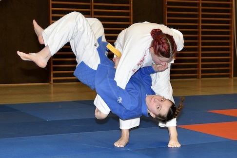 Zwei Judoka f&uuml;hren einen Wurf auf der blauen Judomatte aus, eine K&auml;mpferin befindet sich in der Luft.