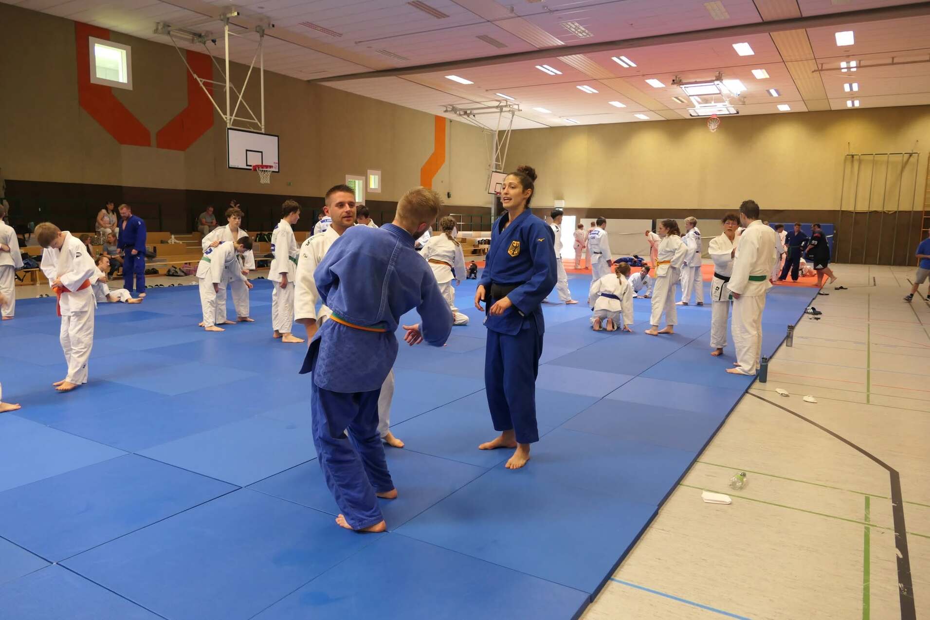 Judo-Training in Sporthalle mit mehreren Kämpfern auf blauen Matten, zwei Athleten unterhalten sich im Vordergrund.