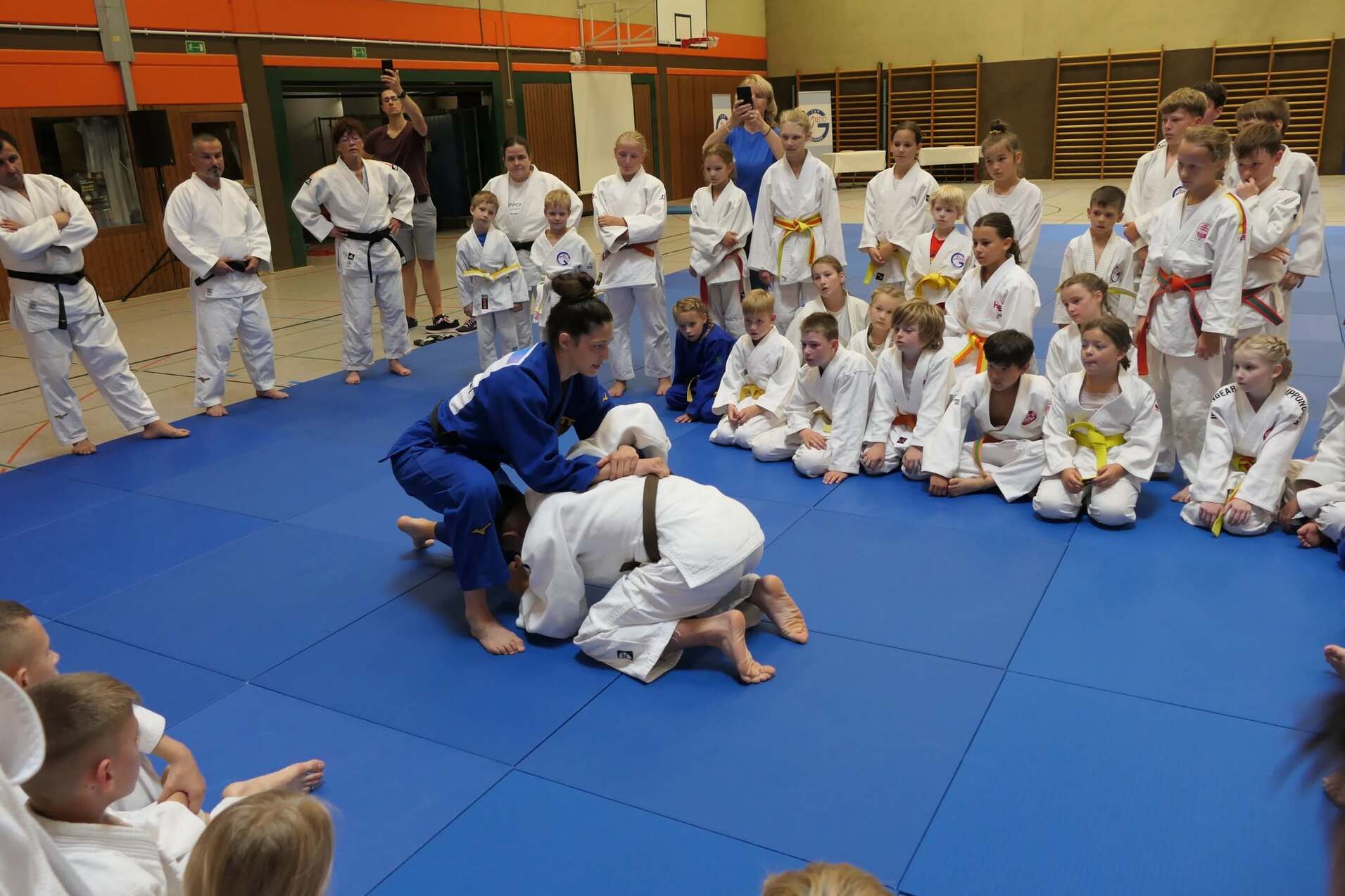 Judo-Training mit Kindern, ein Trainer demonstriert eine Technik auf der blauen Matte, Zuschauer in weißen Gi.