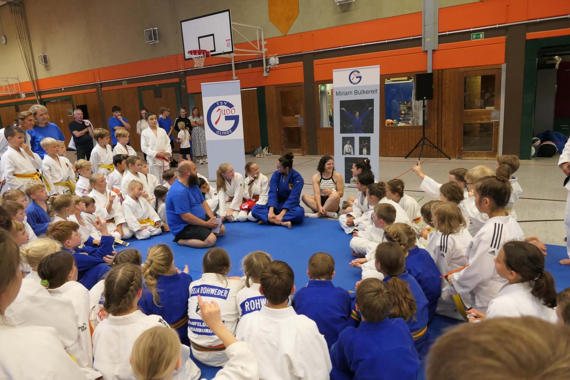 Gruppengespräch mit Kindern in Judokleidung auf einer Matte, umgeben von Trainern in blauen T-Shirts.