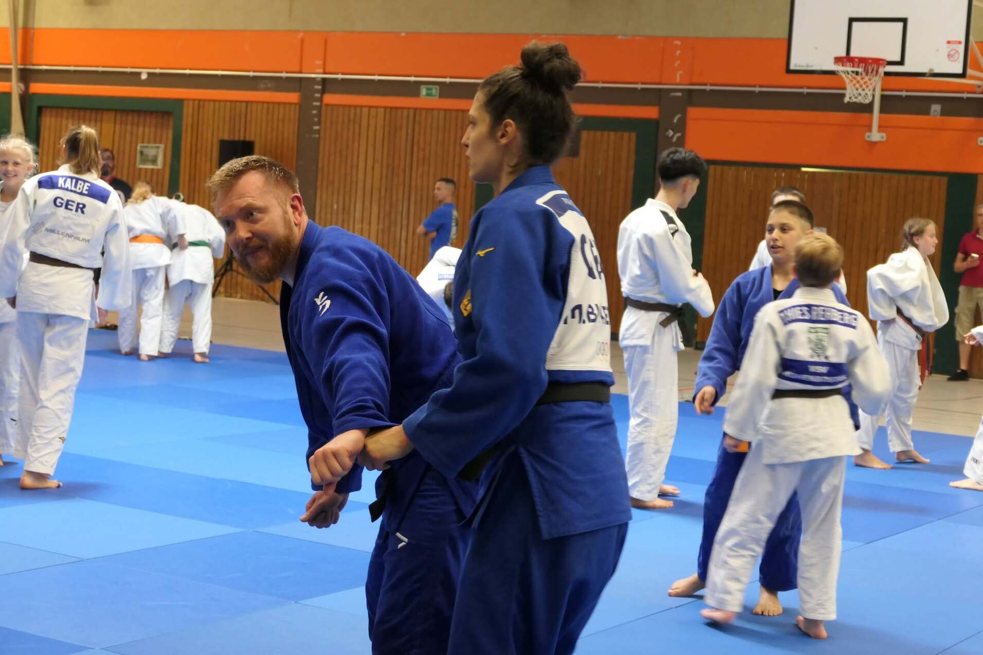 Judo-Training in einer Sporthalle: Zwei Teilnehmer üben einen Wurf, während weitere Judokas im Hintergrund trainieren.