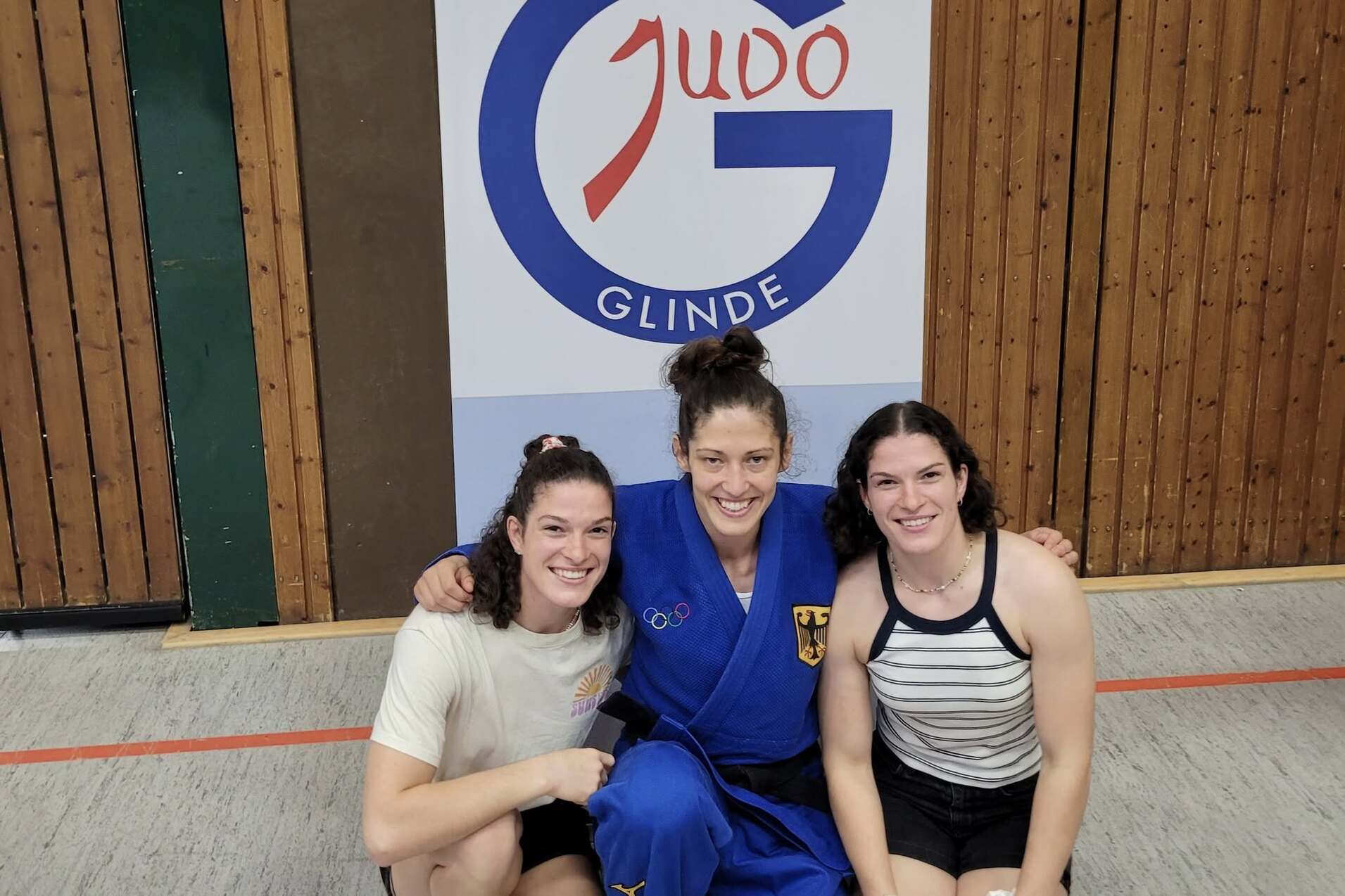 Drei Judo-Athletinnen posieren lächelnd vor einer Wand mit dem Logo des Judo-Clubs Glinde.