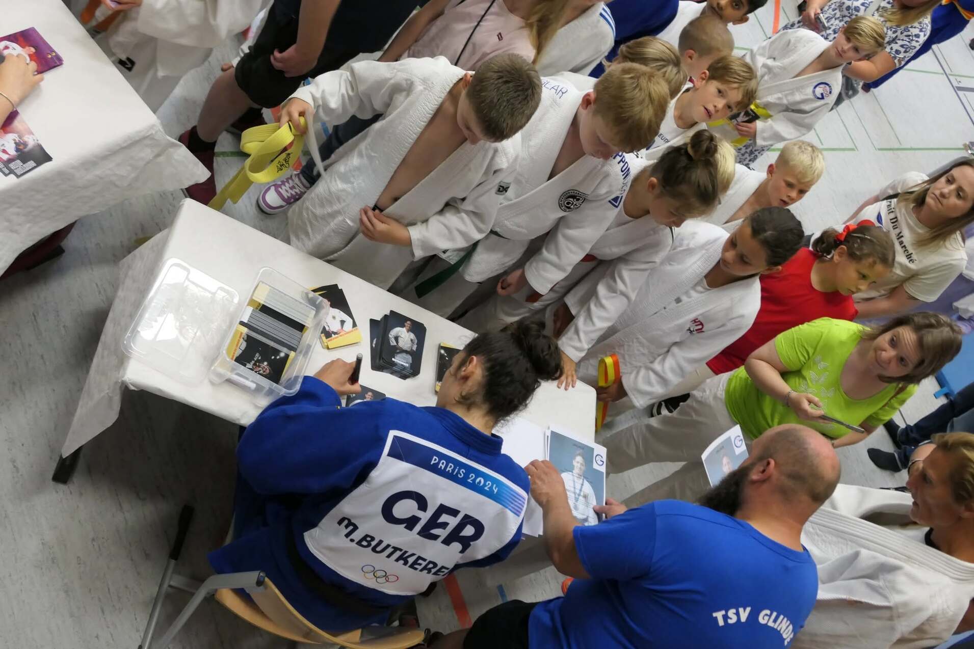 Eine Gruppe von Kindern in Judogi beobachtet einen Judoka beim Signieren von Fotos am Tisch.