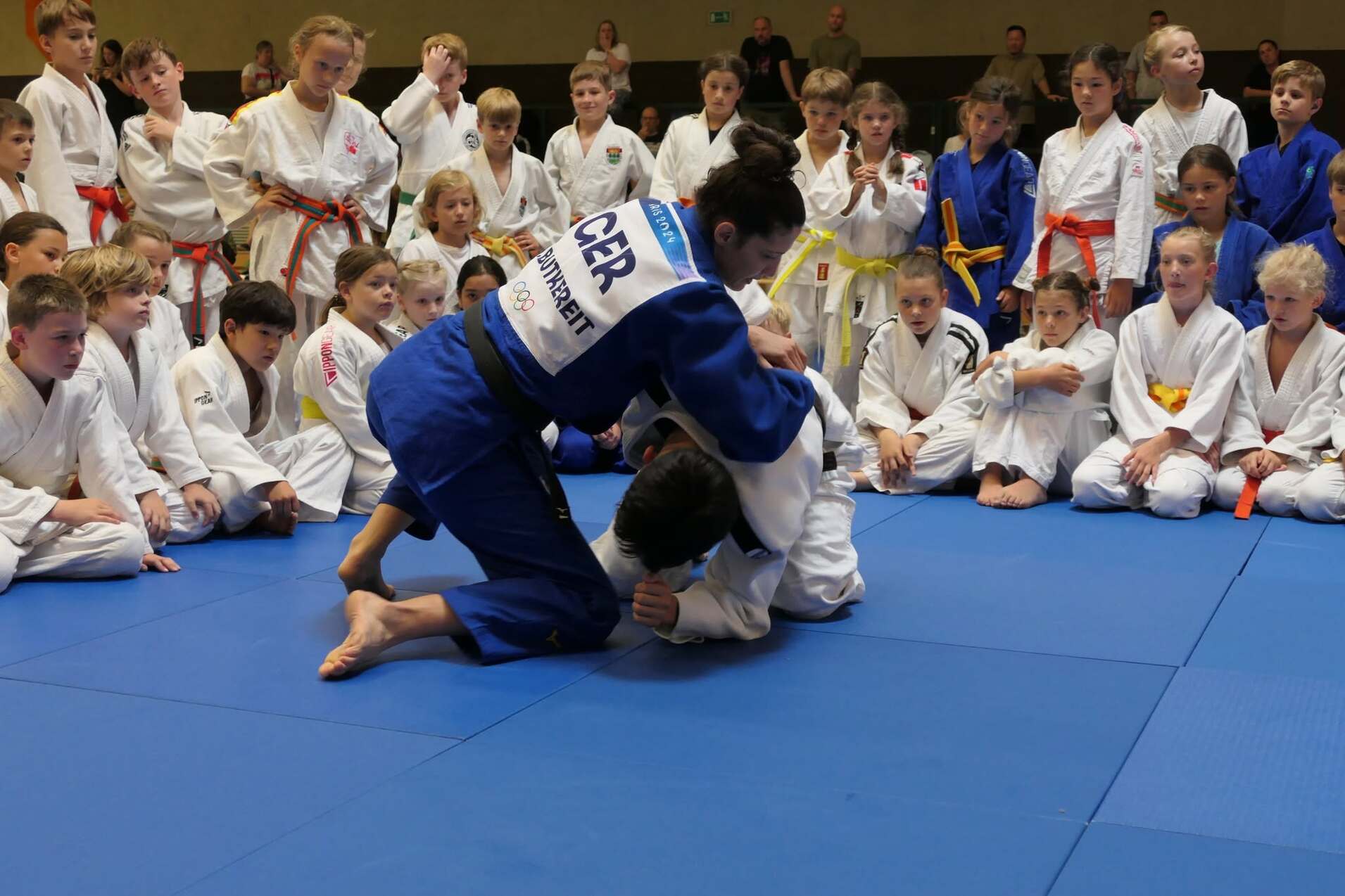 Eine Judo-Praktikantin in blauer Gi führt einen Wurf gegen einen Partner auf einer blauen Judomatte aus, Publikum beobachtet.