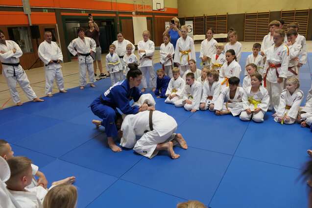 Judo-Training mit Kindern, ein Trainer demonstriert eine Technik auf der blauen Matte, Zuschauer in wei&szlig;en Gi.