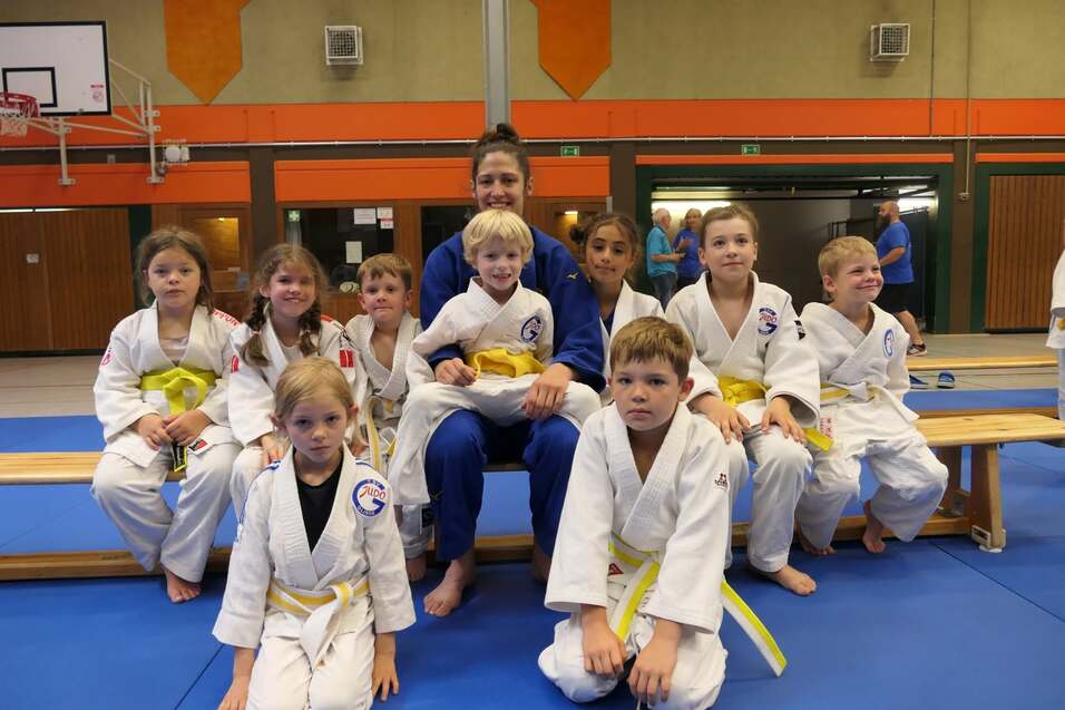 Eine Gruppe von Kindern in wei&szlig;en Judogi mit gelben G&uuml;rteln sitzt und steht um eine Trainerin in blauer Judokleidung.