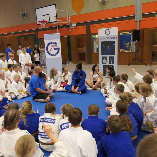 Gruppengespräch mit Kindern in Judokleidung auf einer Matte, umgeben von Trainern in blauen T-Shirts.