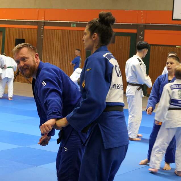 Judo-Training in einer Sporthalle: Zwei Teilnehmer üben einen Wurf, während weitere Judokas im Hintergrund trainieren.