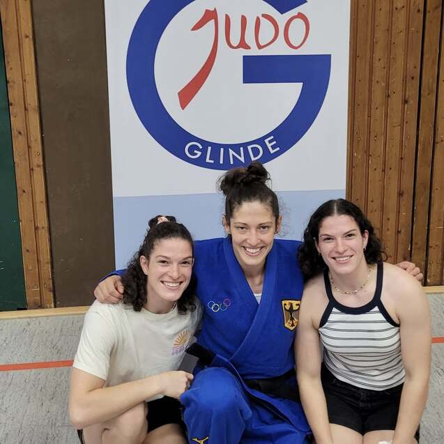 Drei Judo-Athletinnen posieren lächelnd vor einer Wand mit dem Logo des Judo-Clubs Glinde.