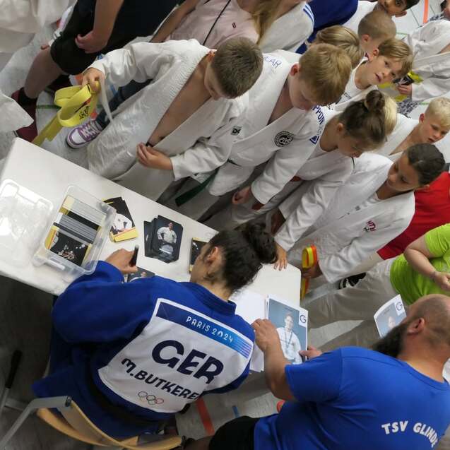 Eine Gruppe von Kindern in Judogi beobachtet einen Judoka beim Signieren von Fotos am Tisch.