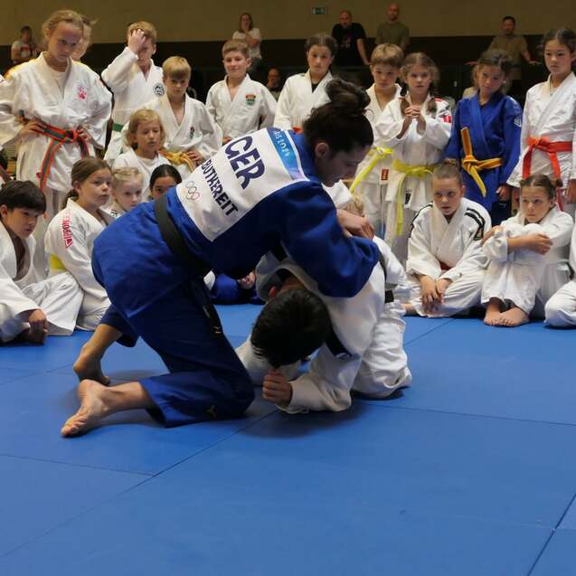 Eine Judo-Praktikantin in blauer Gi führt einen Wurf gegen einen Partner auf einer blauen Judomatte aus, Publikum beobachtet.