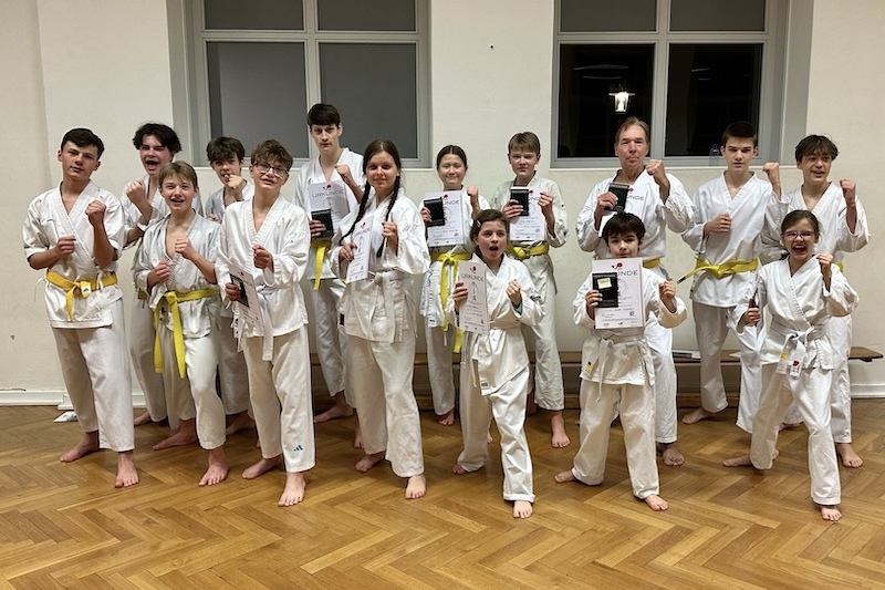 Gruppe von Jugendlichen in Wei&szlig; und Gelb in Karate-Uniformen, posieren stolz mit Urkunden in einem Raum.