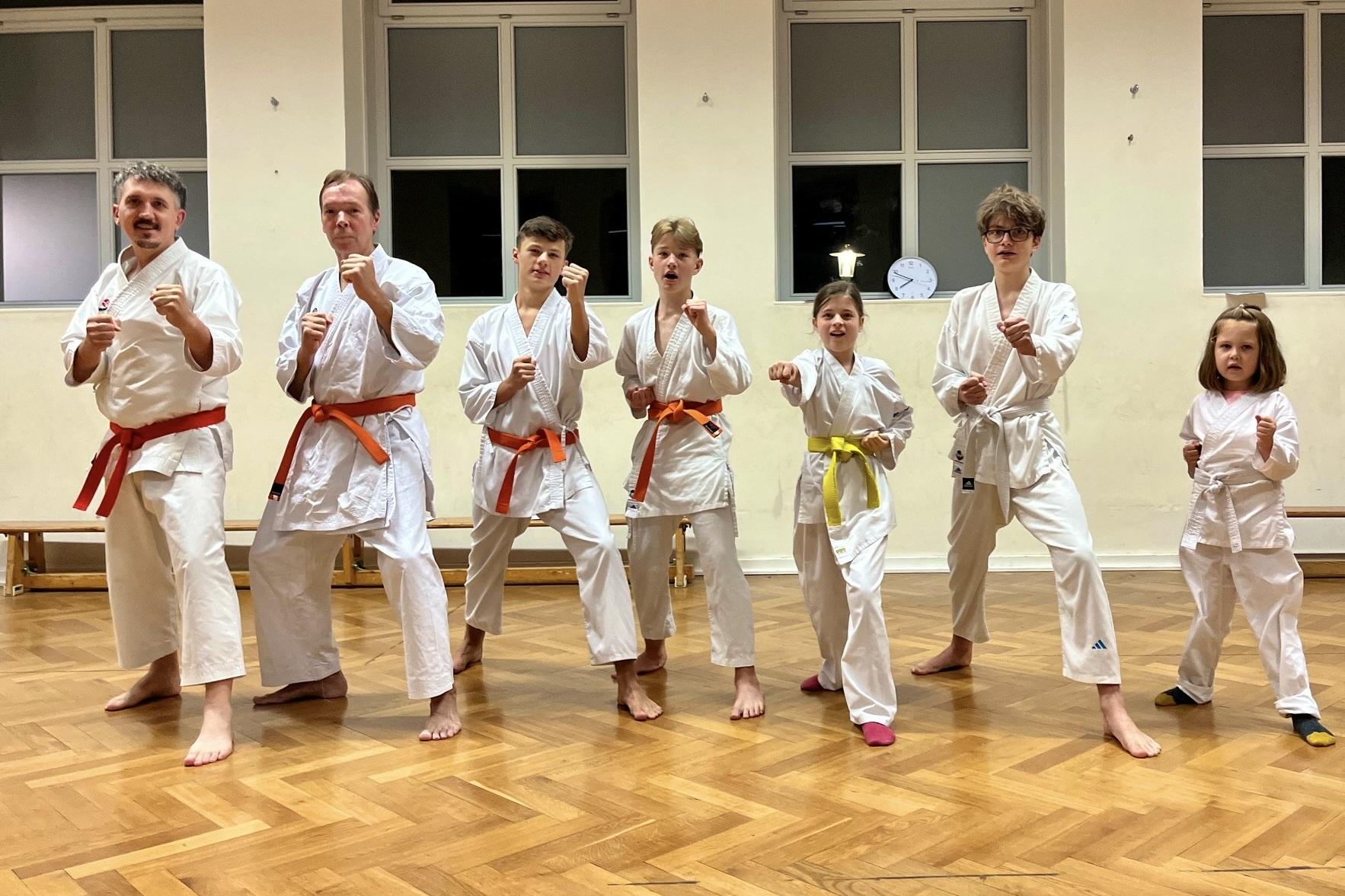 Sechs Personen in Karate-Training, gekleidet in weiße Anzüge mit unterschiedlichen Gürtelfarben, stehen in Kampfstellung.
