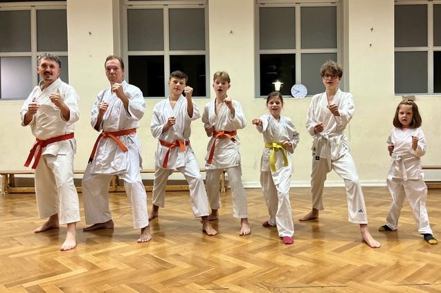 Sechs Personen in Karate-Training, gekleidet in wei&szlig;e Anz&uuml;ge mit unterschiedlichen G&uuml;rtelfarben, stehen in Kampfstellung.