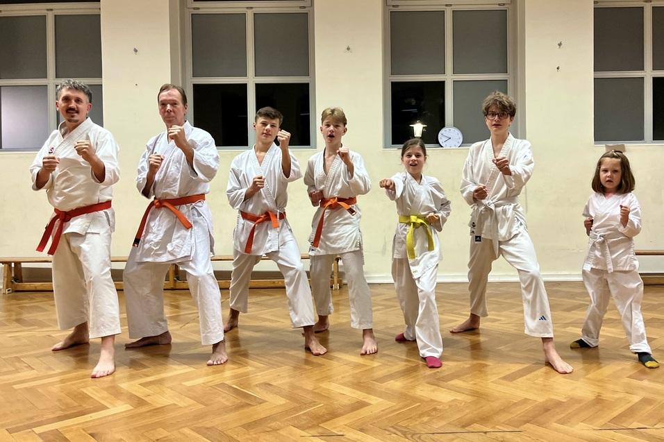 Sechs Personen in Karate-Training, gekleidet in wei&szlig;e Anz&uuml;ge mit unterschiedlichen G&uuml;rtelfarben, stehen in Kampfstellung.