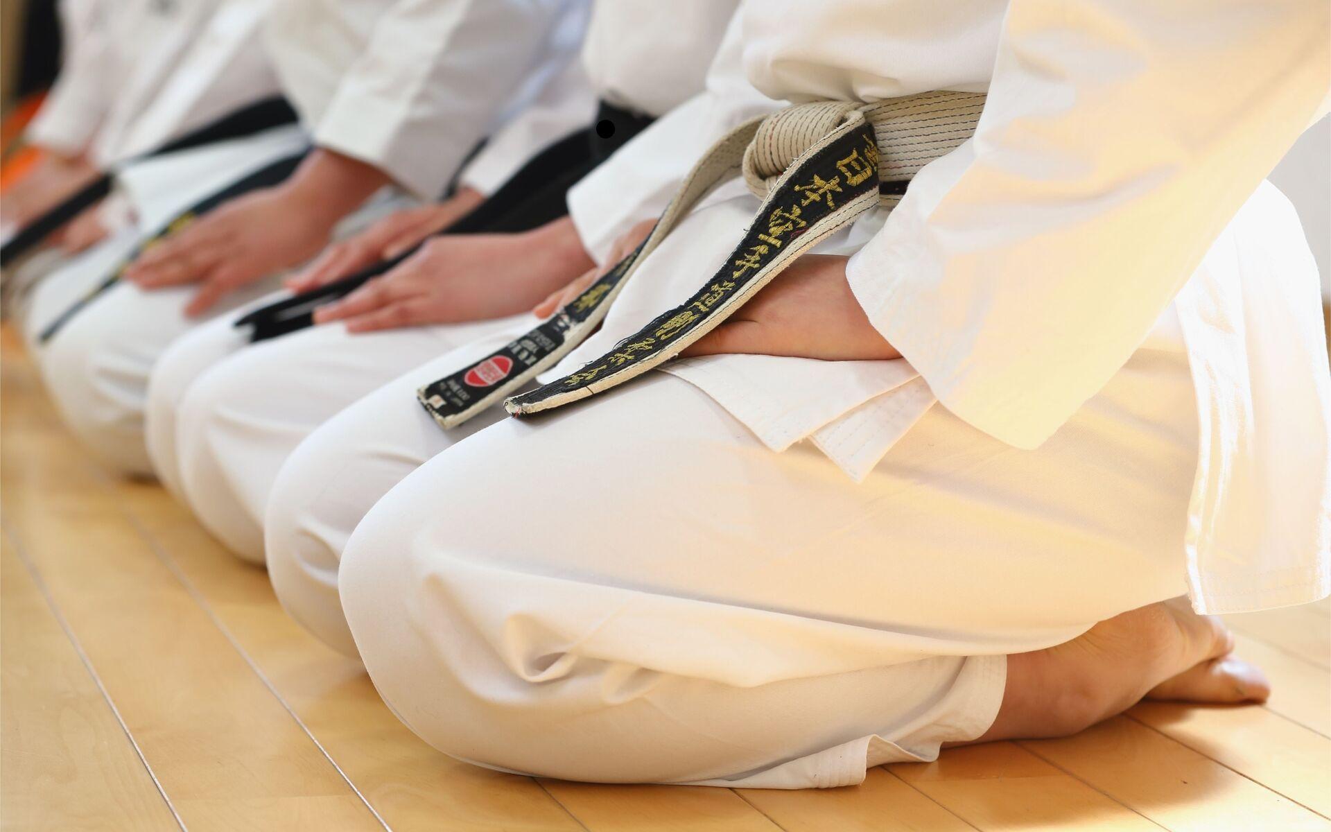 Nahaufnahme von mehreren Karateka im Sitz, tragen weiße Anzüge und schwarze Gürtel, auf einem Holzboden.