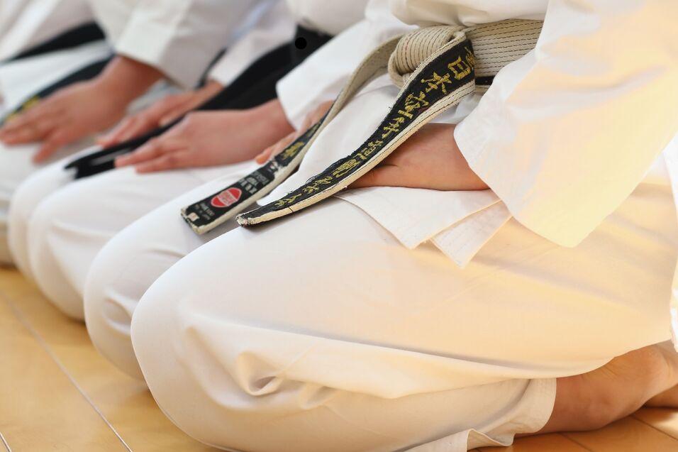 Nahaufnahme von mehreren Karateka im Sitz, tragen weiße Anzüge und schwarze Gürtel, auf einem Holzboden.