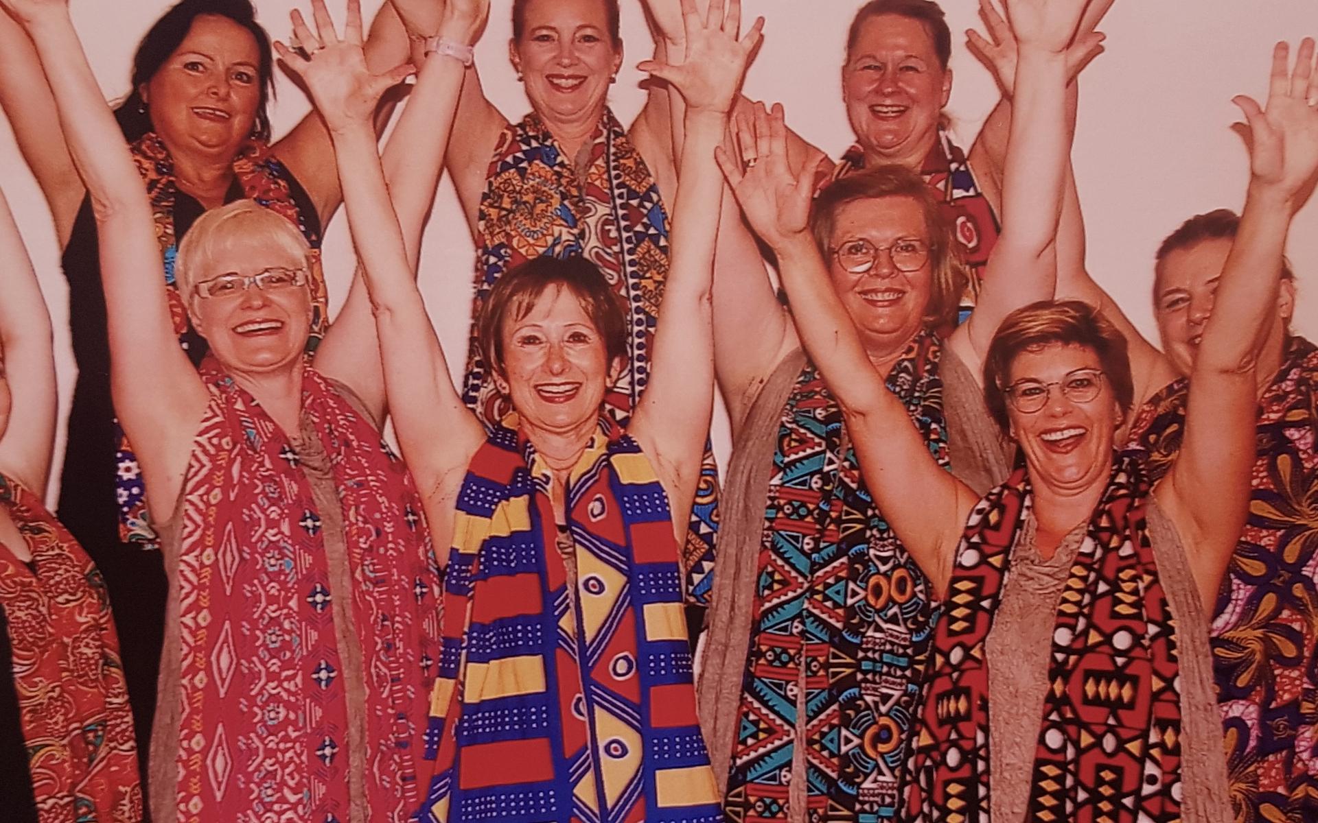 Gruppenshot von neun Personen in bunten Tüchern, die lächeln und die Hände in die Luft strecken.