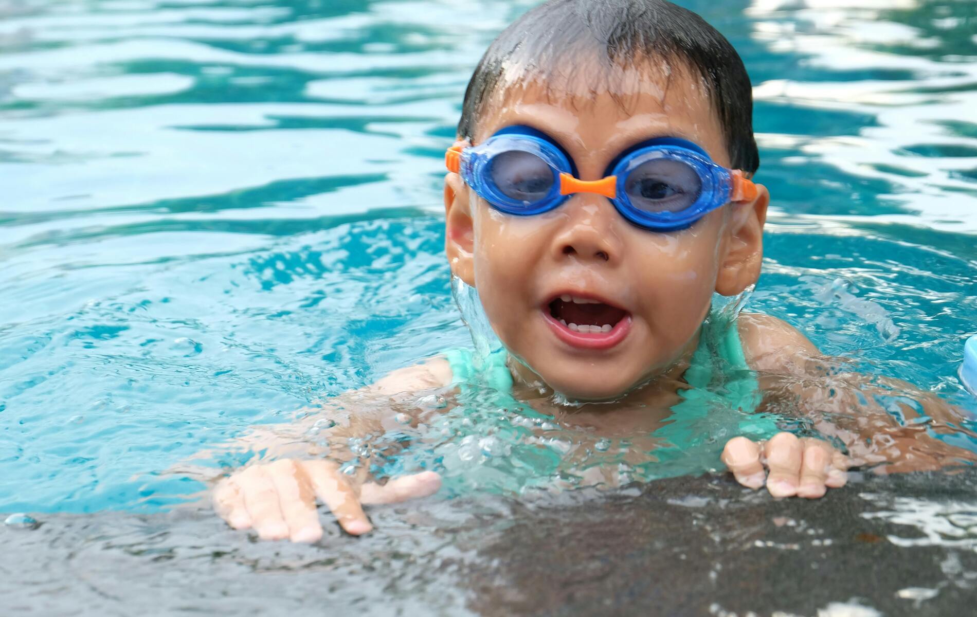 Ein Junge mit blauen Schwimmbrillen schwimmt im Wasser und lächelt, während er am Beckenrand hoch schaut.