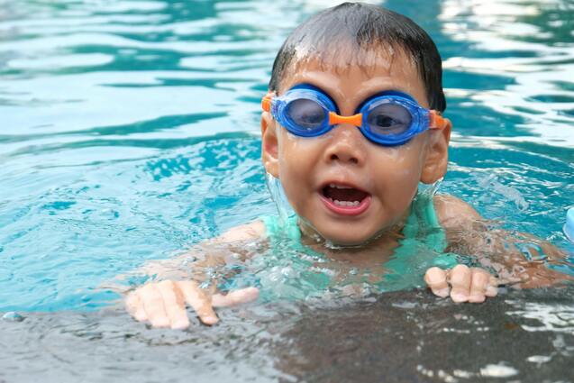 Ein Junge mit blauen Schwimmbrillen schwimmt im Wasser und lächelt, während er am Beckenrand hoch schaut.