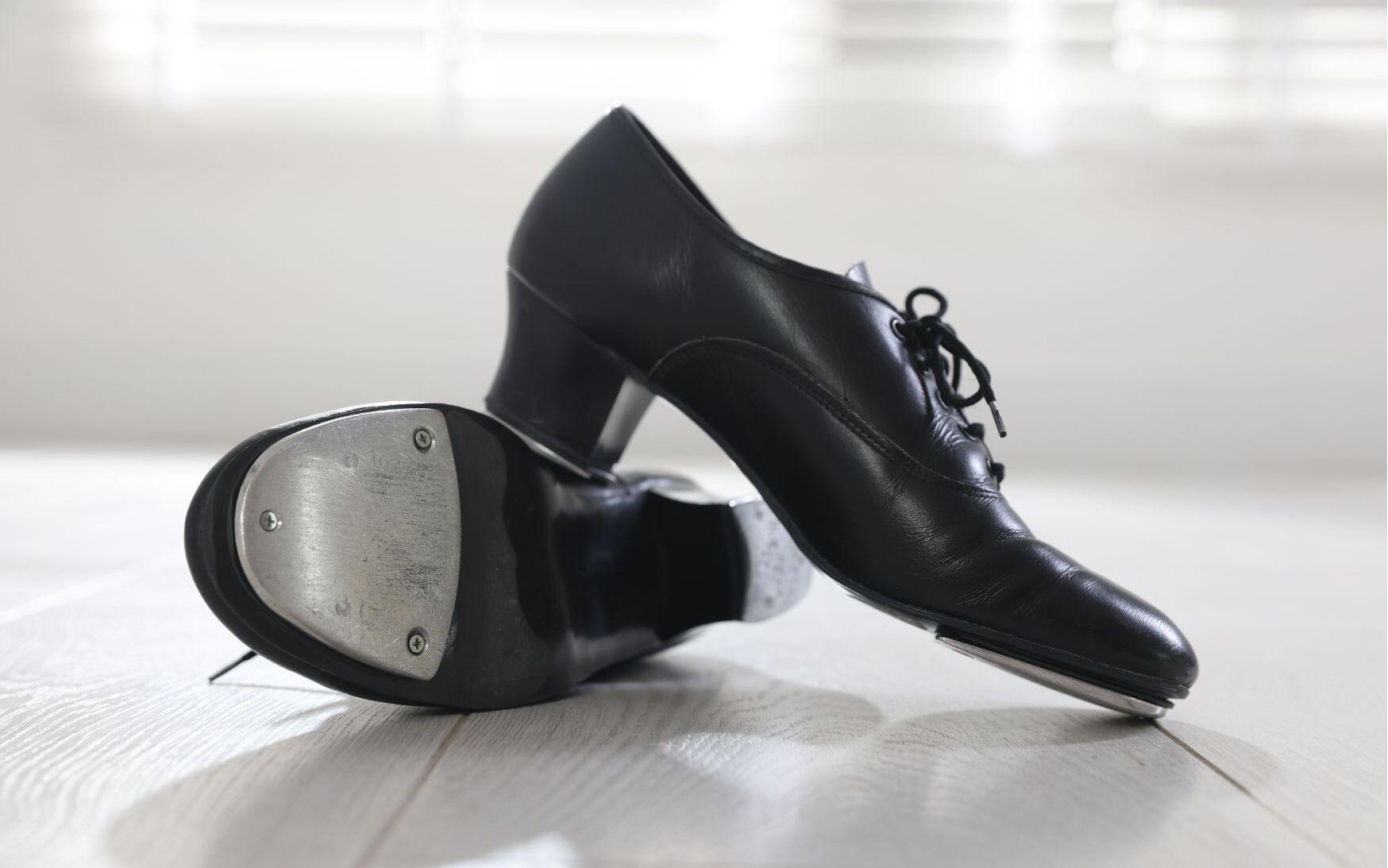 Schwarze Tanzschuhe mit Metalldekoration auf einem hellen Holzfußboden, im Hintergrund sanftes Licht.