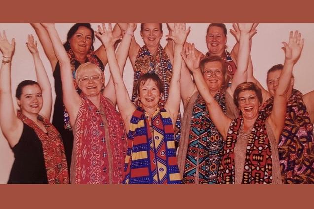 Gruppe von neun Personen in bunten Schals, die fröhlich die Hände heben und in die Kamera lachen.