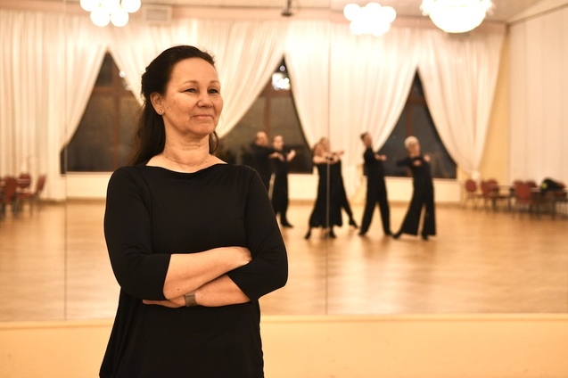 Frau mit verschr&auml;nkten Armen steht vor einem Spiegel in einem Tanzsaal, im Hintergrund tanzen mehrere Personen.