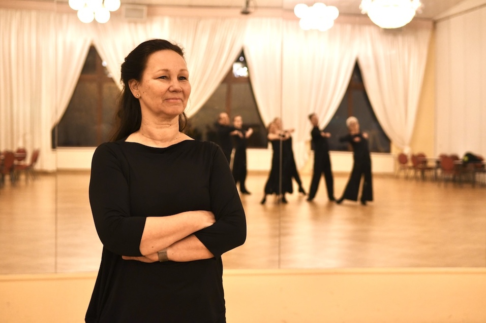 Frau mit verschr&auml;nkten Armen steht vor einem Spiegel in einem Tanzsaal, im Hintergrund tanzen mehrere Personen.