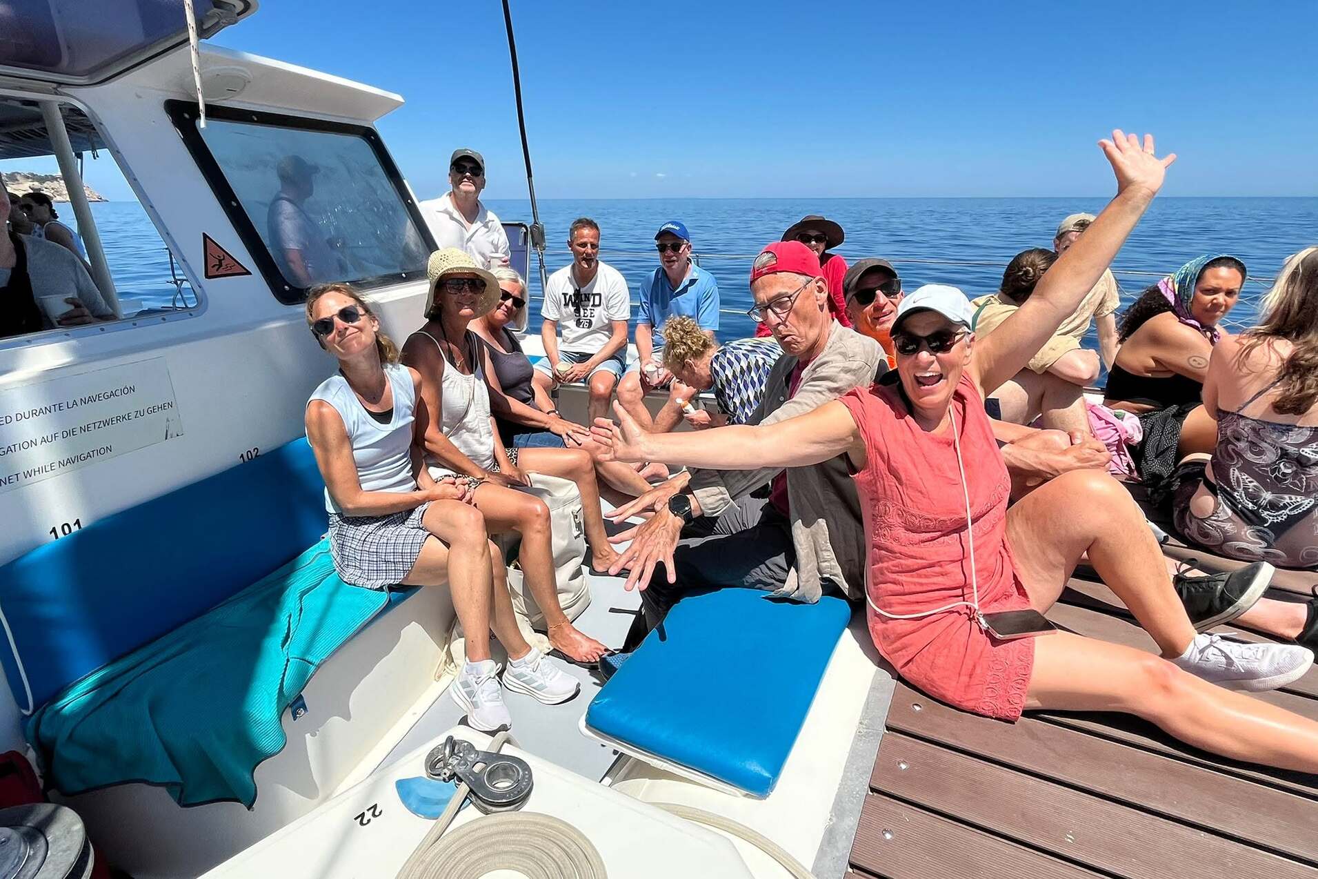 Gruppe von Personen entspannt auf einem Boot, einige winken, w&auml;hrend andere sich unterhalten, bei sonnigem Wetter.