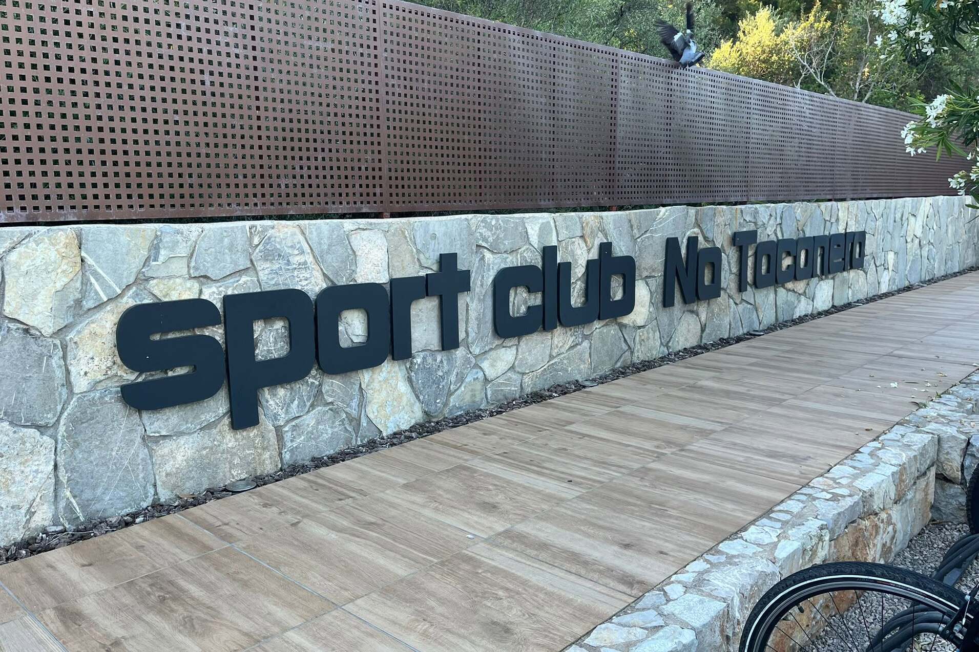 Schild des Sportclubs &bdquo;Na Tonnara&ldquo; an einer Steinmauer, umgeben von gr&uuml;ner Natur und Holzweg.