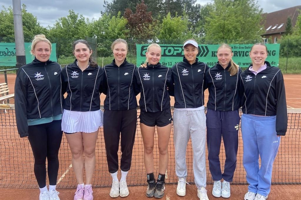 Sieben Tennisspielerinnen in Sportjacken stehen lächelnd auf einem Sandplatz vor einem Netz.