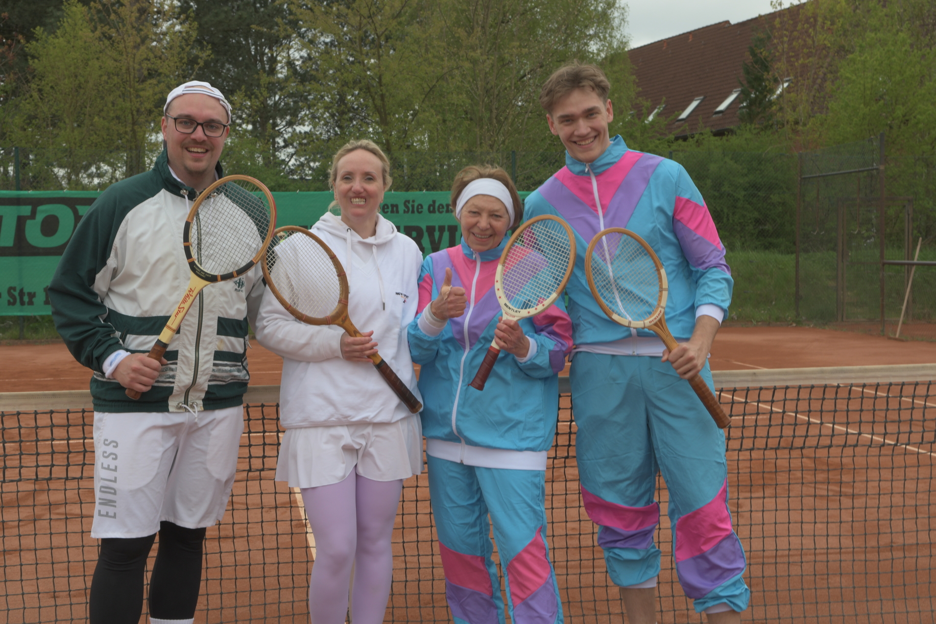 Vier Personen in sportlicher Kleidung halten Tennis-Schl&auml;ger auf einem Tennisplatz mit Sandbelag.