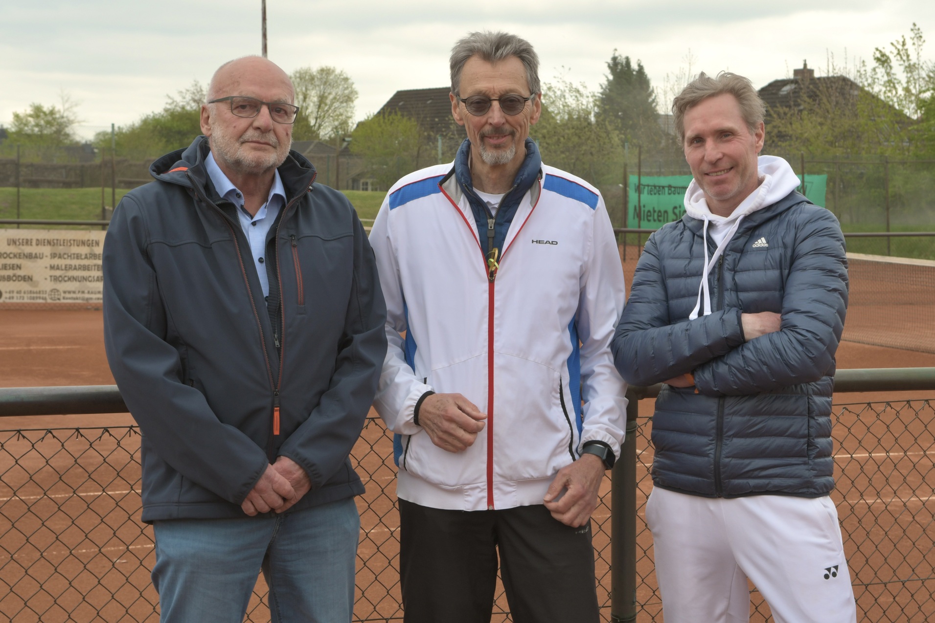 Drei M&auml;nner stehen l&auml;ssig vor einem Tennisplatz, im Hintergrund sind B&auml;ume und eine Tennisanlage sichtbar.