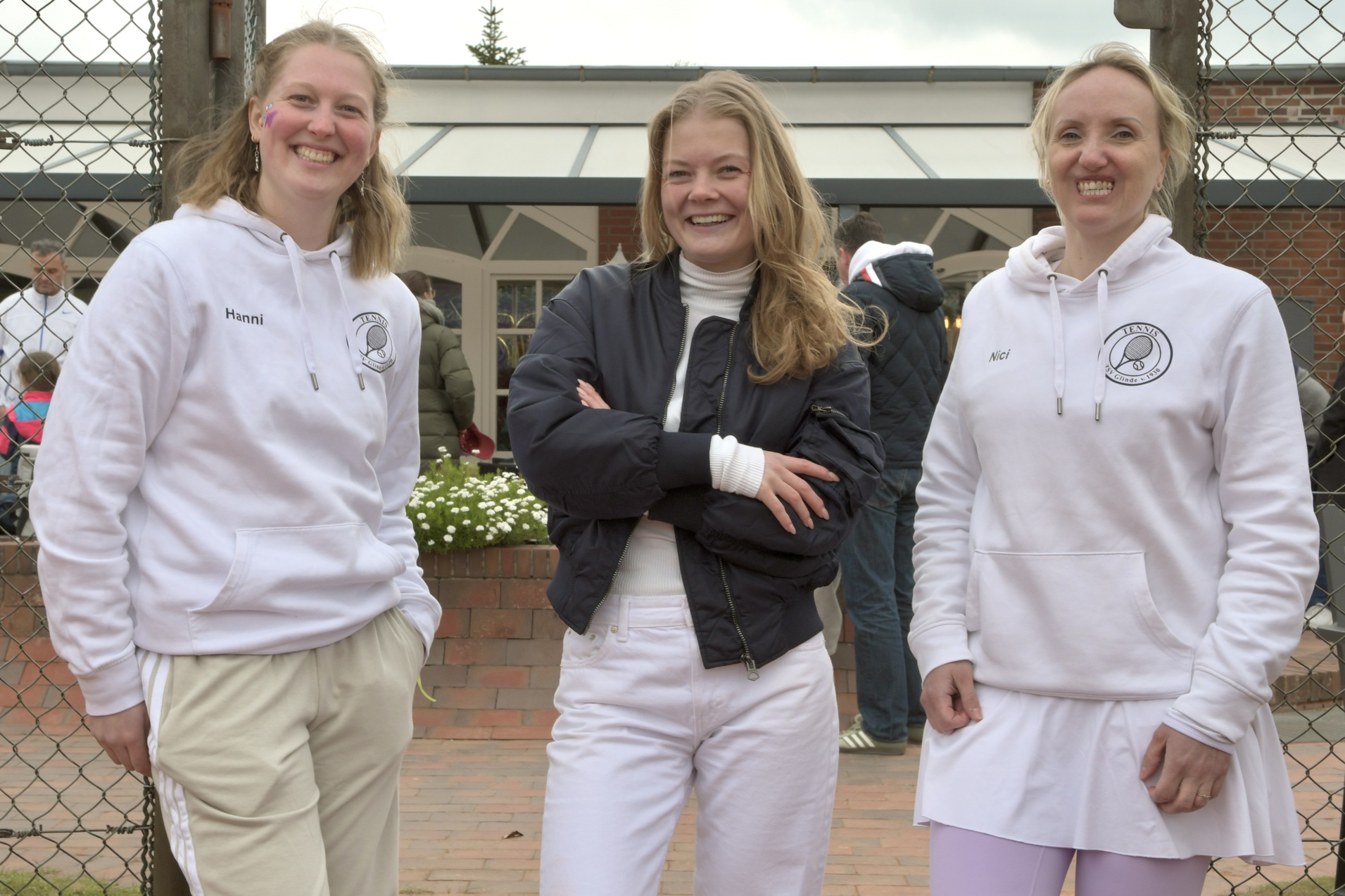 Drei Frauen in Sportkleidung stehen l&auml;chelnd vor einem Tennisplatz mit einem Zaun im Hintergrund.