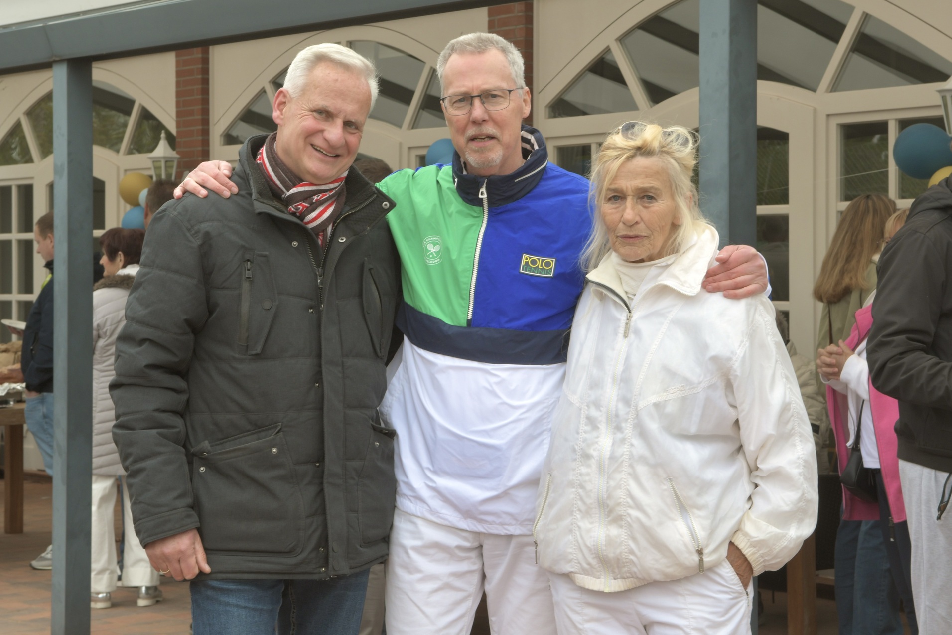 Drei Personen stehen l&auml;chelnd zusammen, eine Frau in wei&szlig;er Sportkleidung, zwei M&auml;nner, einer davon in einer bunten Jacke.
