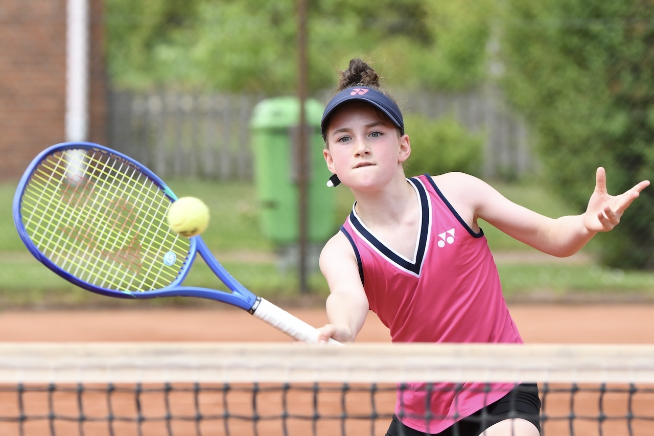 Junge Tennis-Spielerin in pinkem Trikot schl&auml;gt einen Ball mit dem Schl&auml;ger auf einem Tennisplatz.