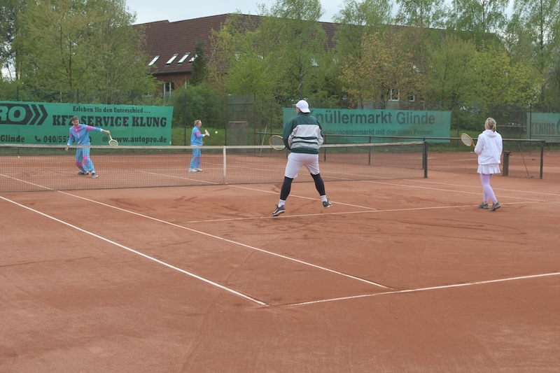 Drei Tennisspieler auf einem sandigen Platz, zwei in blauen Outfits, einer in grauer Jacke und schwarzer Hose, spielen ein Match.