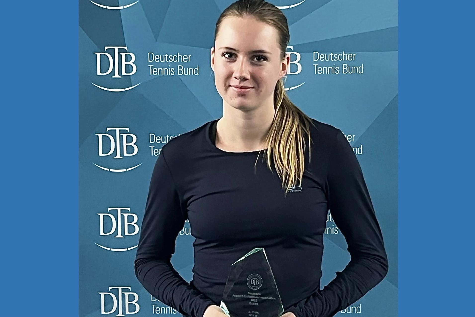 Junge Frau in sportlicher Kleidung hält einen Pokal vor einem Hintergrund mit dem Logo des Deutschen Tennis Bundes.