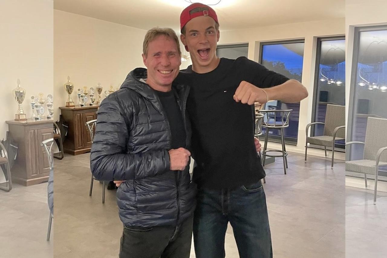 Zwei junge Männer umarmen sich, lächelnd vor einem Raum mit Pokalen und Stühlen im Hintergrund.