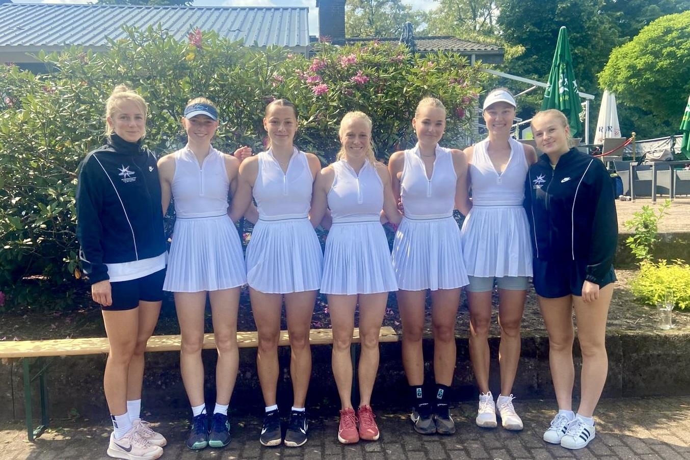 Gruppe von sieben Tennis-Spielerinnen in weißen Dresses, lächelnd vor einer grünen Kulisse im Freien.