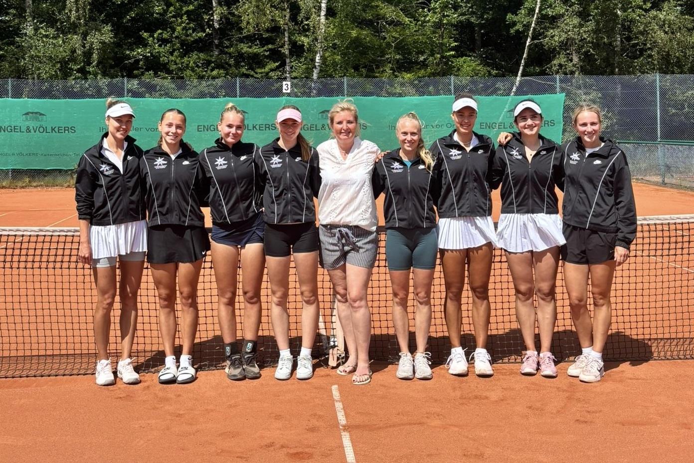 Acht weibliche Tennis-Spielerinnen und eine Trainerin posieren in Teamkleidung vor einem Tennisnetz auf einem Sandplatz.