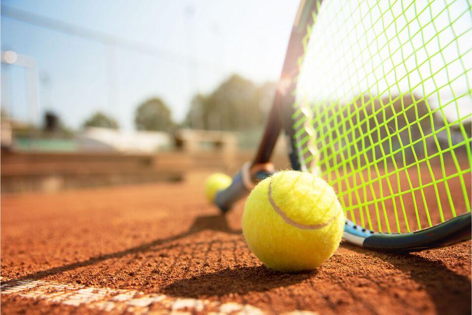 Nahaufnahme eines tennisballs auf einem roten Sandplatz, neben einem Tennisnetz und Schläger.