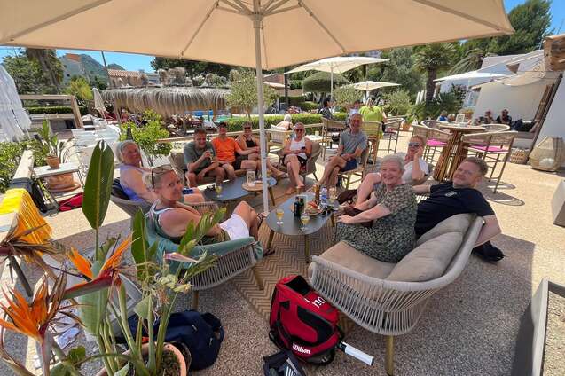 Gruppe von acht Personen sitzt unter einem Sonnenschirm in einem modernen Caf&eacute; mit Pflanzen und stilvollen M&ouml;beln.