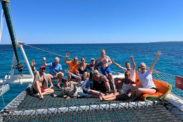 Gruppe von 15 Personen auf einem Katamaran-Netz, l&auml;chelnd und winkend, mit blauem Meer im Hintergrund.