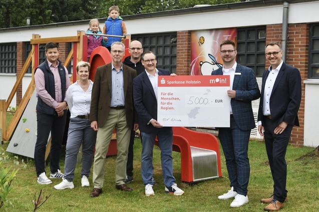 Gruppenszene mit sechs Erwachsenen und zwei Kindern, die einen gro&szlig;en Scheck &uuml;ber 5000 Euro f&uuml;r ein Projekt pr&auml;sentieren.