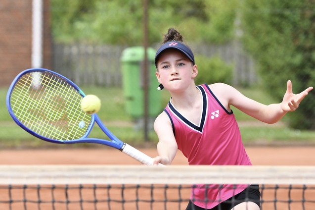 Junge Tennis-Spielerin in pinkem Trikot schl&auml;gt einen Ball mit dem Schl&auml;ger auf einem Tennisplatz.